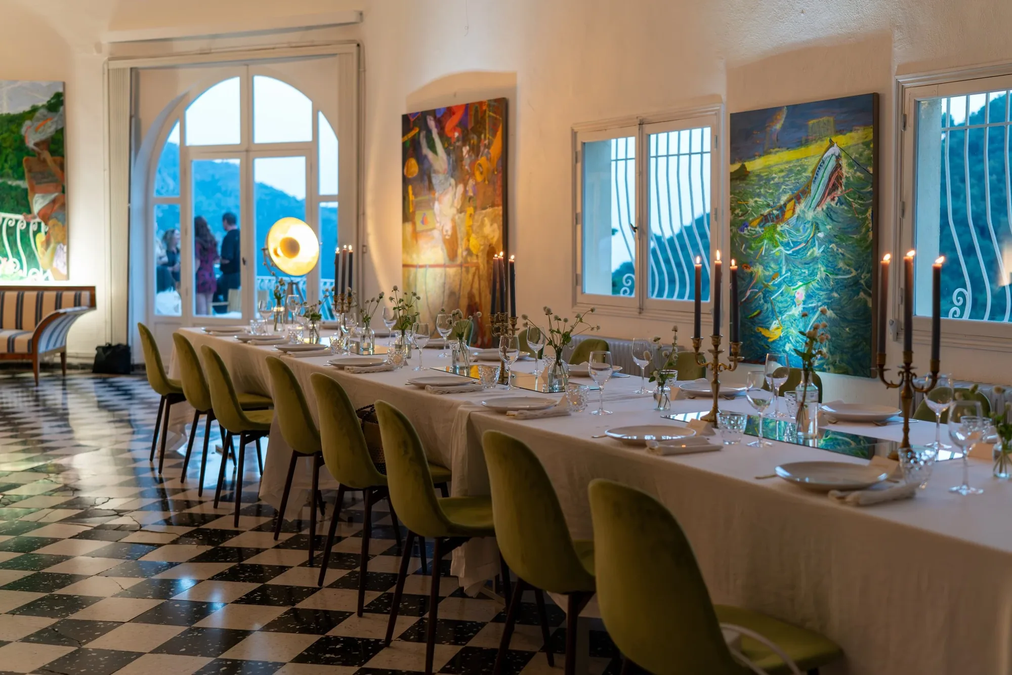 A long candlelit dinner table set in the salon at Castel Caramel, with paintings on the walls and guests on the terrace
