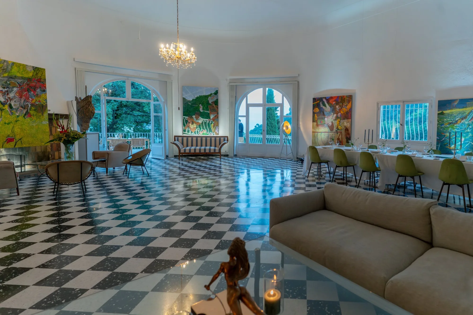 The salon at Castel Caramel at dusk, with paintings by Henry Curchod on the walls, a chandelier and the dinner table laid